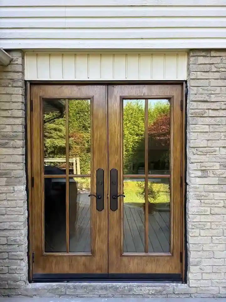 Custom wood-grain fiberglass double doors with divided glass panels and bronze handle sets with thumb latches on a brick and vinyl home