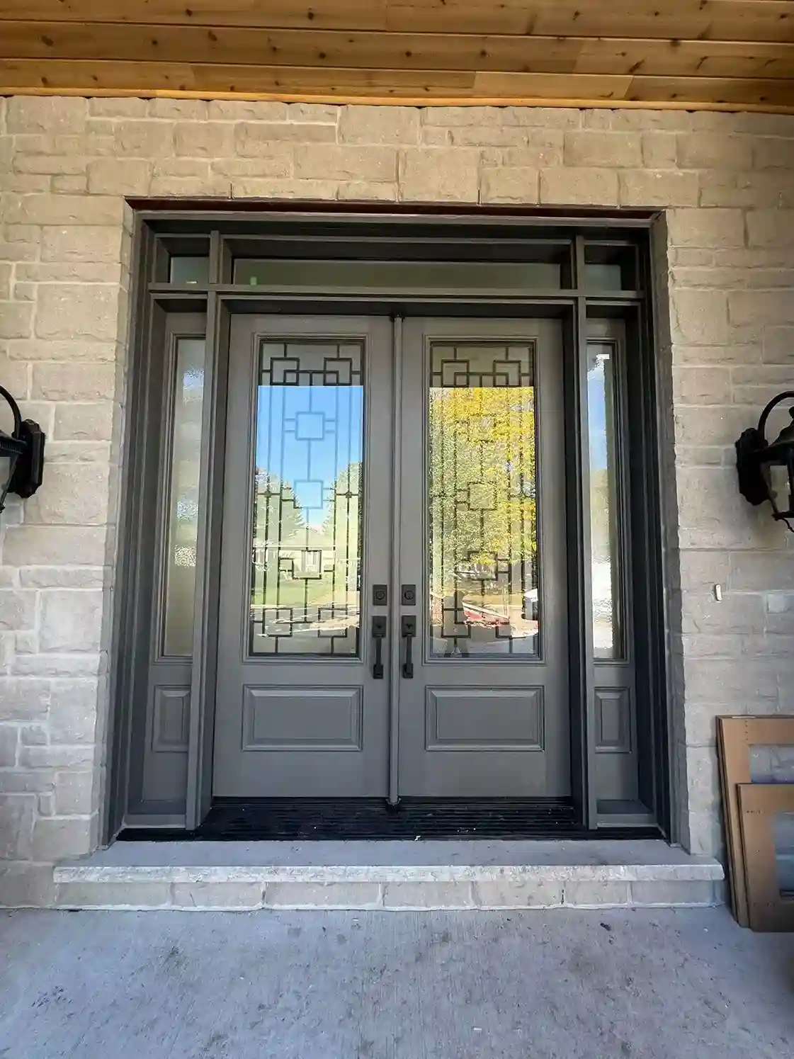Grey fiberglass double doors with decorative glass, sidelights and a transom, set in a stone wall under a covered porch